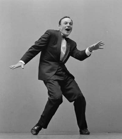 a black and white photo of a man wearing a suit, tap dancing against a solid gray backdrop. the man is middance move, leaning forward and smiling, with his arms outstretched. he is wearing a tuxedo with a bow tie and has his hair slicked back. the lighting is typical of a studio setting, with even illumination and no harsh shadows.