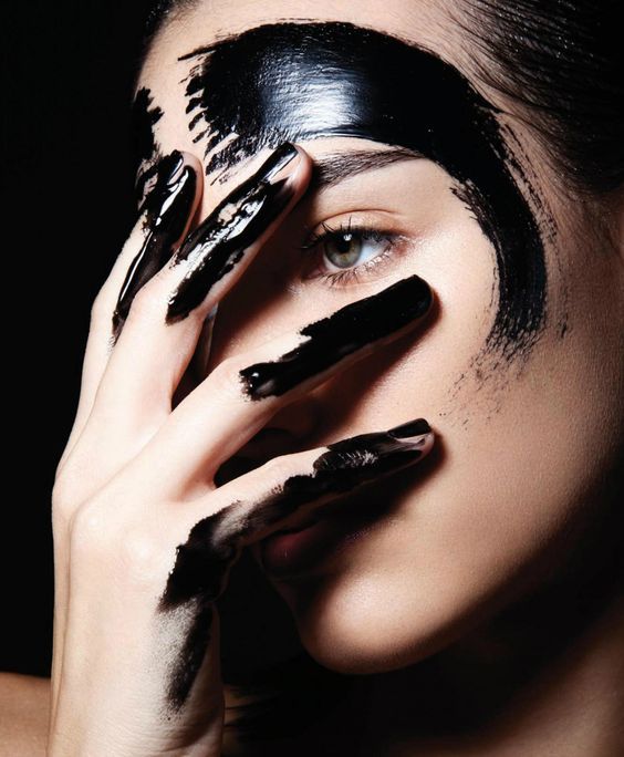 a closeup portrait of a woman with black paint on her face and hands. her right hand is covering her left eye and a portion of her face, with paint splattered over her fingers. the paint creates a dramatic effect, highlighting her features. the woman's expression is serene and mysterious. the background is dark, creating a stark contrast to the vibrant colors of the paint. this image is a beautiful example of creative makeup and photography.