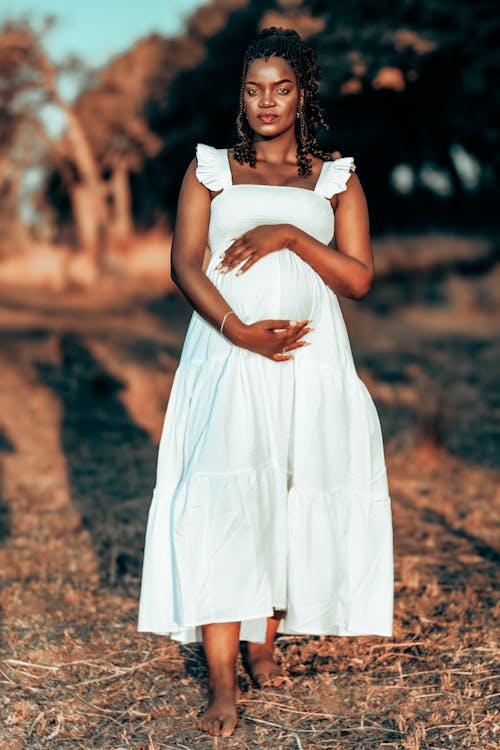 a pregnant black woman stands in a field, barefoot, wearing a flowing white dress.  the setting appears to be outdoors in nature, with sunlight illuminating the scene. she has her hands gently resting on her pregnant belly, radiating a serene and peaceful expression.  her hair is styled in braids. the overall mood is one of joy and anticipation of motherhood.