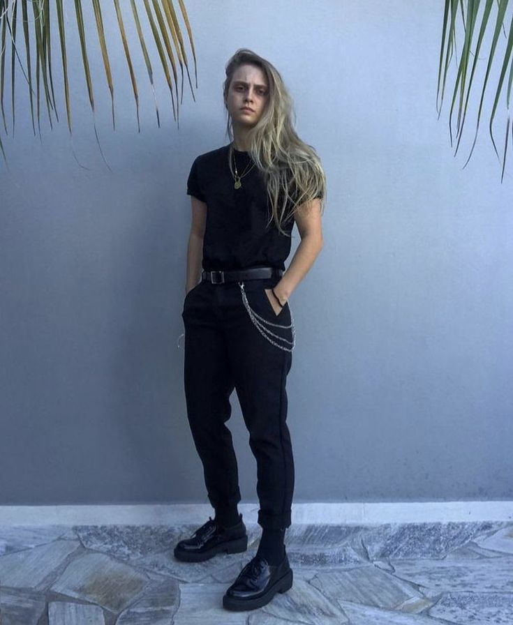 the image shows a young person with long blonde hair wearing an allblack outfit.  the outfit consists of a simple black tshirt, black pants cuffed at the ankles, black oxford shoes, and a chain belt. the person is standing against a light blue wall with palm leaves framing the sides of the image. the overall style is minimalist and androgynous, leaning towards a slightly dark or edgy aesthetic.  the pose is casual and relaxed, with hands in pockets. the background is simple, allowing the clothing and the person's style to be the focal point.