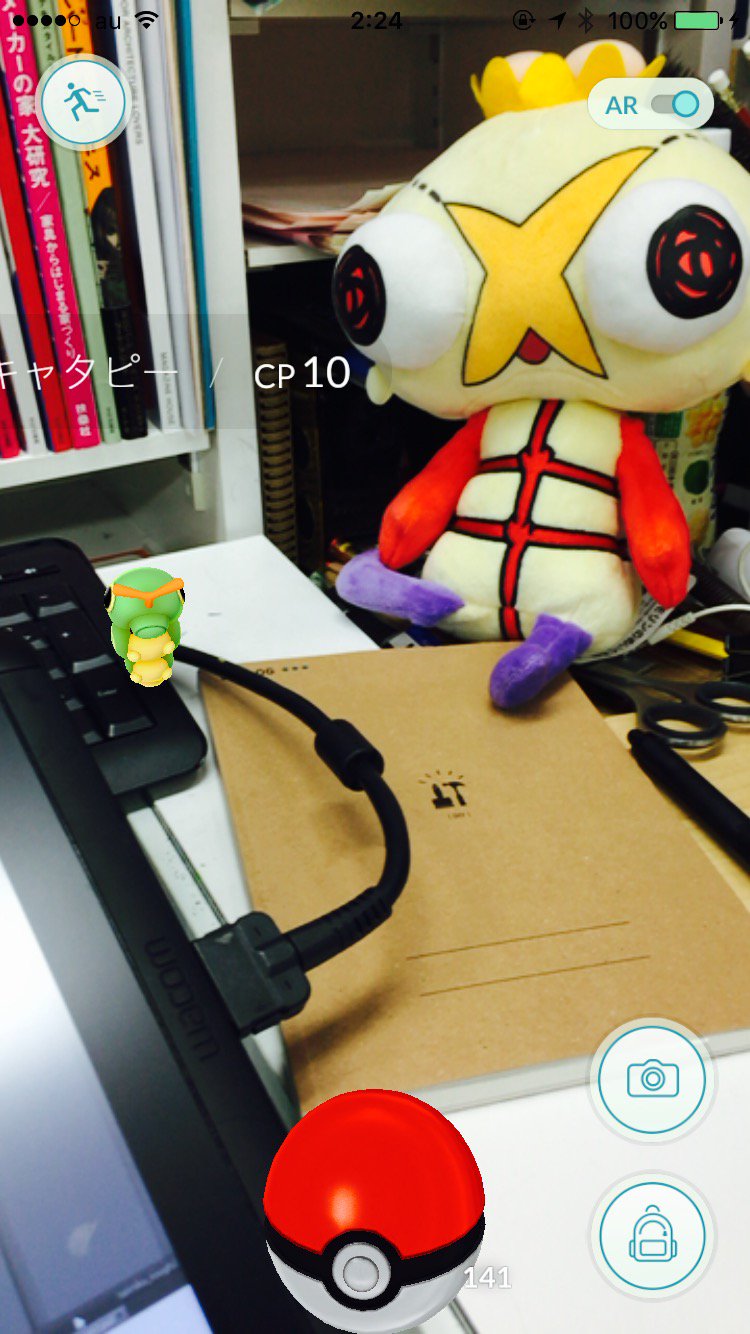 a screenshot of the pokemon go app open on an iphone, with a squirtle pokemon on a tablet. there are books on a shelf and a stuffed toy in the background.  there is a notebook, a pen, and a tablet on the desk.