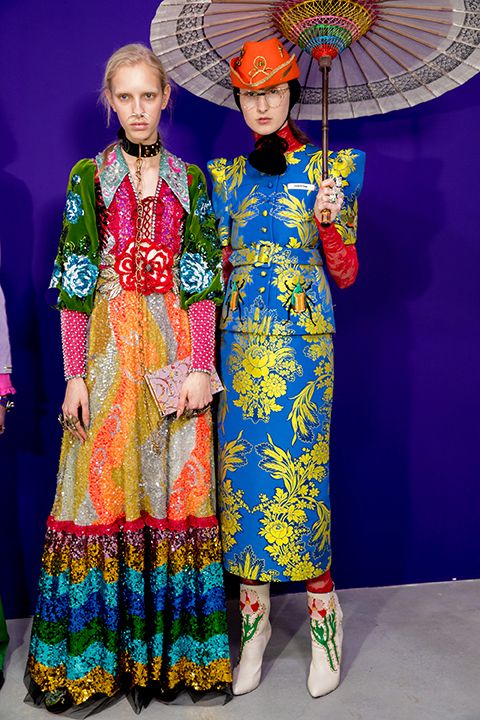 two female models are showcasing vibrant and elaborate gucci outfits on a purple background. the model on the left wears a multicolored sequined dress with layers of rainbow hues, adorned with floral embroidery and velvet accents. she accessorizes with a black choker necklace and carries a pale pink clutch. the model on the right sports a blue brocade suit with a yellow floral pattern, paired with a red cowboy hat, white embroidered boots and long red gloves. she holds an antiquestyle patterned umbrella.  both outfits are richly detailed and exemplify gucci's bold aesthetic. the image is a professional shot, likely from a fashion show.