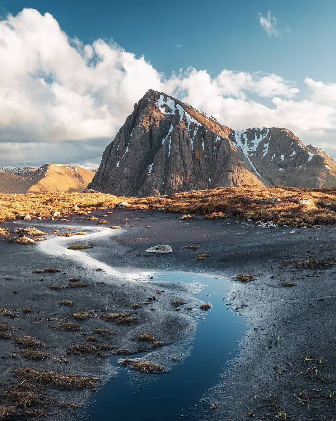 a landscape featuring a mountain under a sky with scattered clouds. a stream meanders through a foreground of dark, textured ground, reflecting the sky. patches of snow cling to the mountain's rugged slopes. the area is surrounded by hills and moorland.