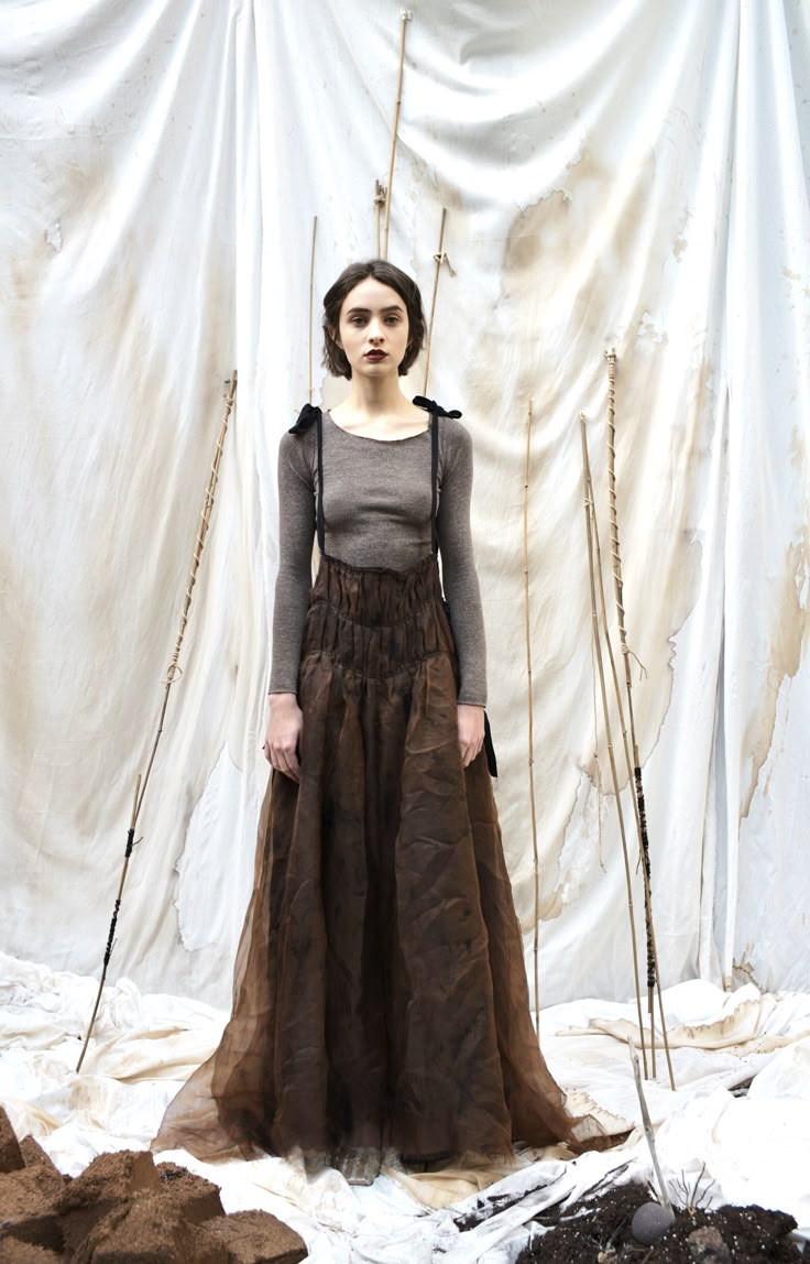 a woman stands facing forward, wearing a longsleeved grey top and a long brown dress with straps and ribbons at the shoulders. she has short dark hair and dark lipstick. the background is a wrinkled white fabric with brown stains and thin sticks leaning against it. the ground is a mix of white fabric and dark soil with scattered objects.