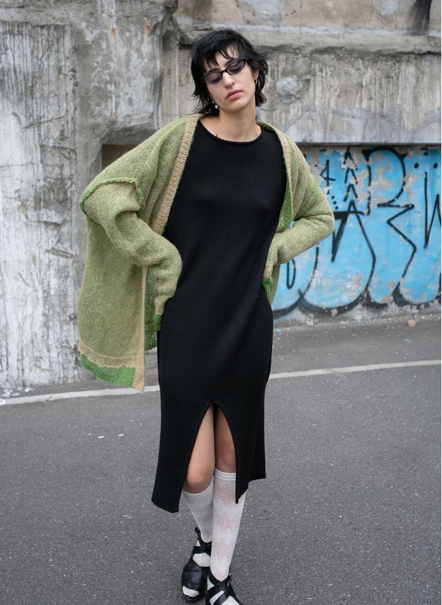 a young woman stands on a city street, posing for a fashion photograph. she's wearing a black midi dress with a front slit, a loosefitting green and beige cardigan, white ankle socks, and black sandals. her hair is short and dark, and she's wearing eyeglasses. the background includes a graffiticovered wall. the overall style is urban and slightly grunge.