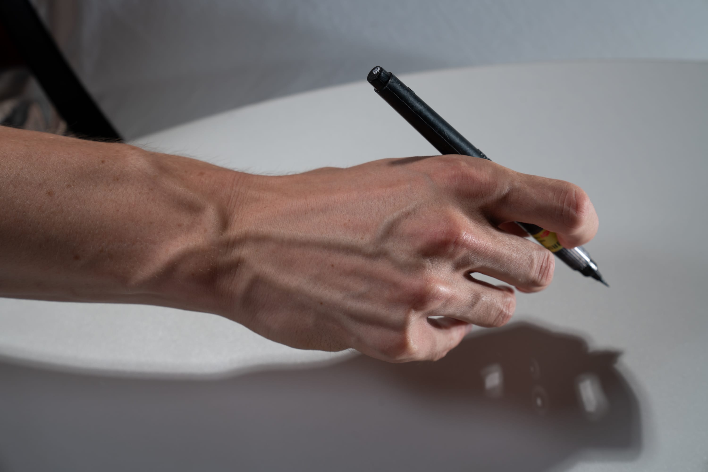 a man's hand is holding a black pen, poised above a white surface. the hand is illuminated by natural light, creating a high contrast image that highlights the veins in the hand and the shadows cast by the pen. the image suggests creativity, writing, or drawing.
