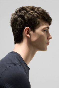 a young man with short, brown hair is captured in a side profile portrait. the lighting is soft and natural, highlighting his defined jawline and features. he wears a simple dark blue tshirt and his expression is neutral. the overall aesthetic suggests a fashion or beauty shoot.