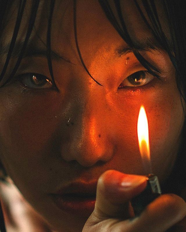 closeup portrait of a woman holding a lit lighter near her face. her eyes are wide and intense, and a single tear is visible. the flame provides illumination, highlighting the skin texture and creating a dramatic effect. she has dark, wet hair framing her face.