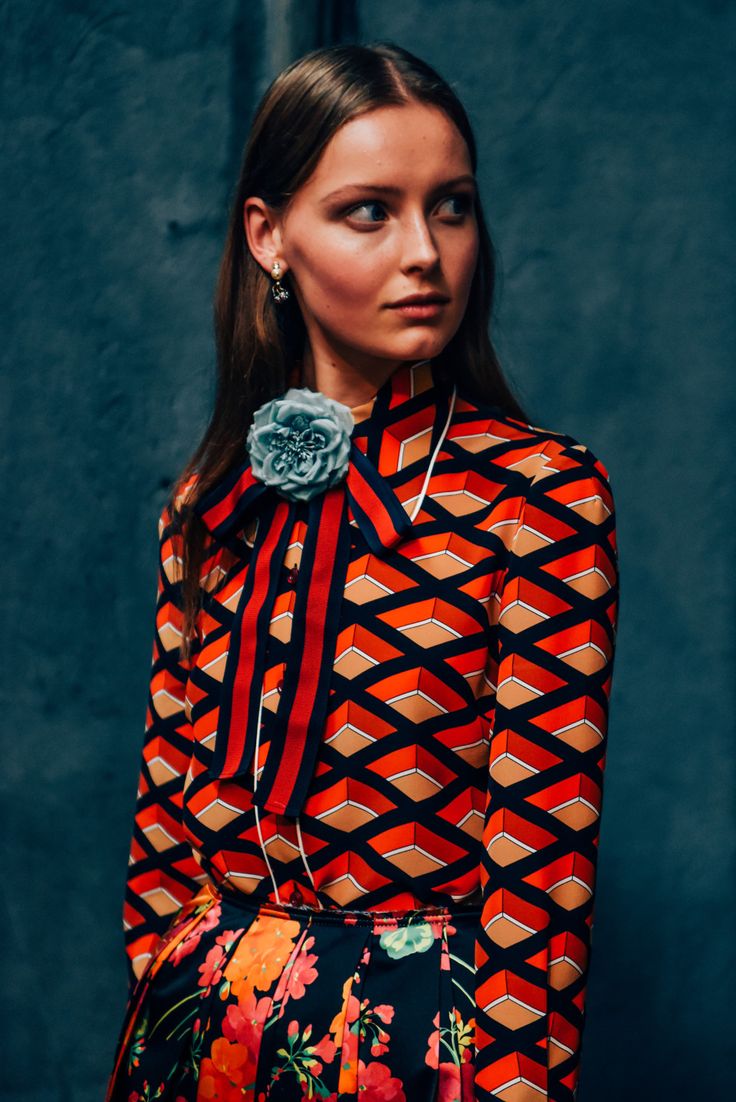 closeup shot of a fashion model wearing a stylish outfit. the model is a young woman with shoulderlength brown hair, wearing a red and black geometric print blouse with a teal flower brooch and red and black ribbon bow. she also wears a black floral print skirt and delicate earrings. the background is a dark teal, creating a striking contrast with the vibrant colors of her clothing. the overall style is elegant and trendy, suggesting an autumn or winter fashion collection. the image is professionally shot, highlighting the details of the clothing and the model's features.