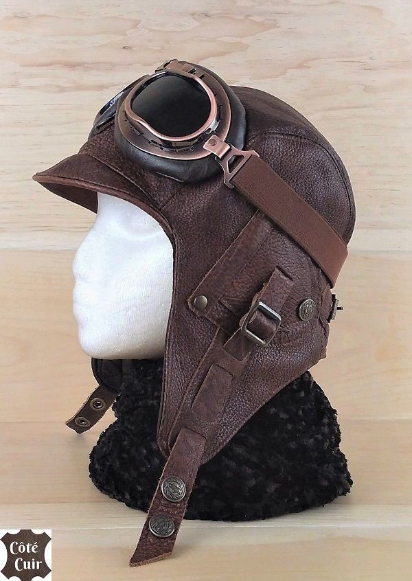 a brown leather aviator hat with goggles on top is displayed on a white head mannequin. the hat has a small brim and ear flaps with straps that buckle. the goggles have dark lenses and coppercolored frames. the mannequin is placed on a wooden surface with a dark, possibly fur, covering around its neck. a logo for "côté cuir" is visible in the bottom left corner.