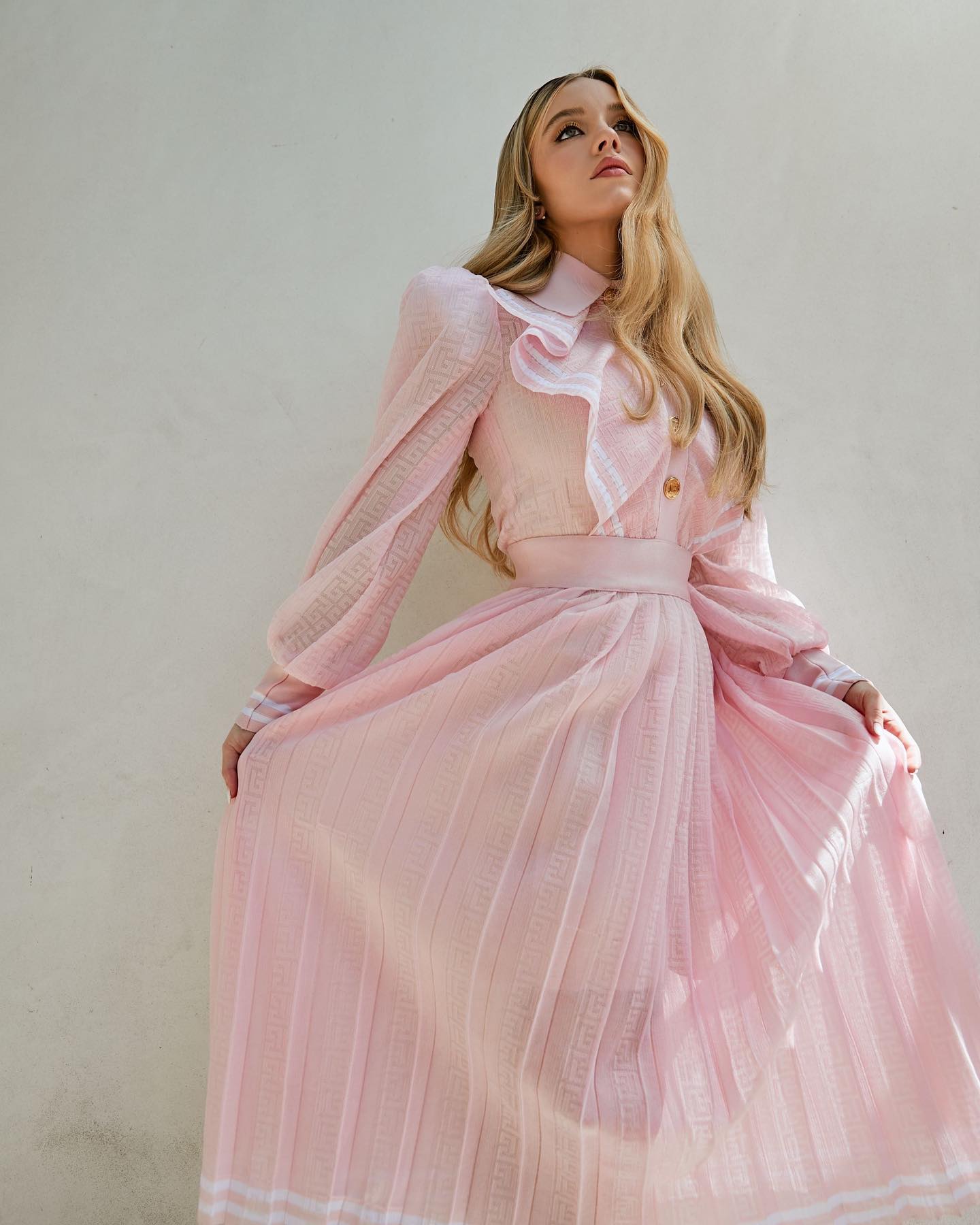 a young blonde woman in a pretty, long, pleated pink dress with ruffle collar is posing against a white background.  the dress is sheer and has a flowing, graceful look. the model is looking up and slightly to her right. the image is welllit and has a soft, feminine feel.