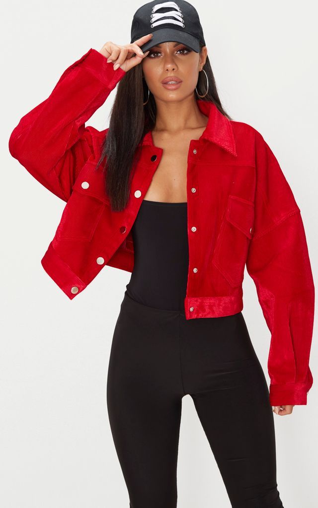a woman is shown modeling a red corduroy cropped jacket. she is also wearing black leggings and a black baseball cap with white laces.  the background is plain white. the image is a fashion photograph showcasing the jacket.