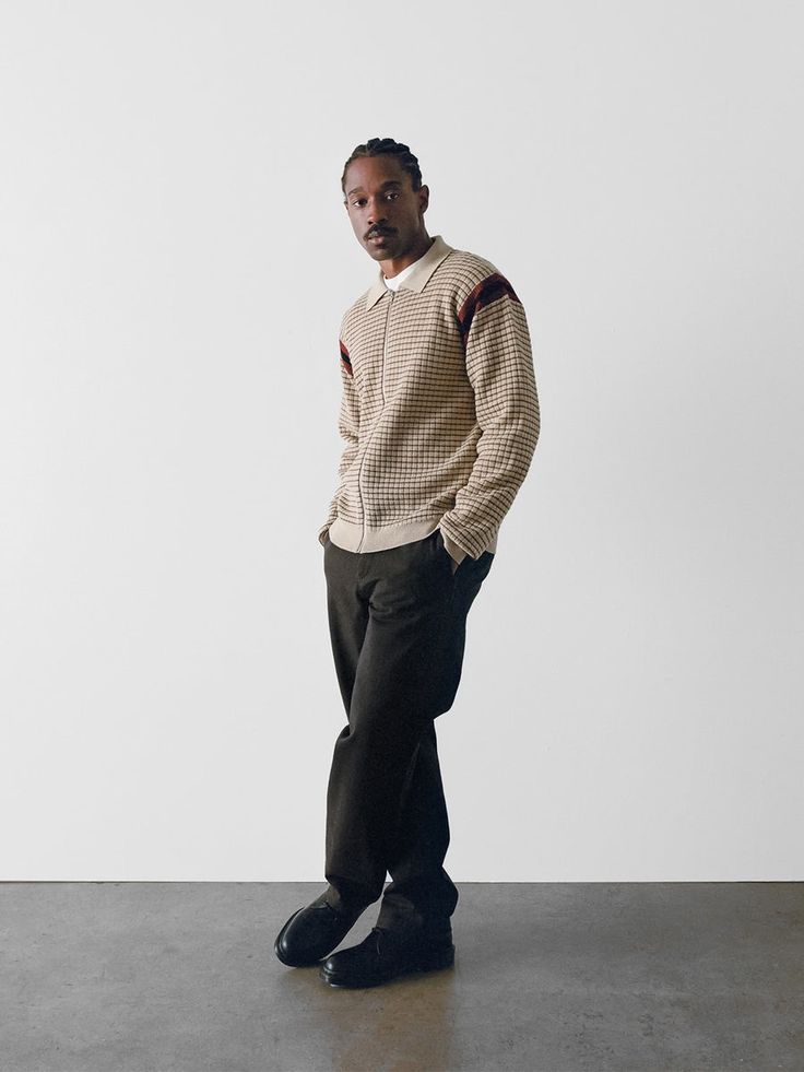 a fullbody shot of a man standing against a plain white background. he's wearing a beige checkered zipup cardigan with brown accents on the sleeves, dark brown trousers, and black loafers. his pose is relaxed, hands in his pockets, and he's looking directly at the camera with a serious expression. the image has a minimalist aesthetic, with the focus entirely on the man and his clothing. the lighting is even, and the background is unblemished, emphasizing the details of the outfit.