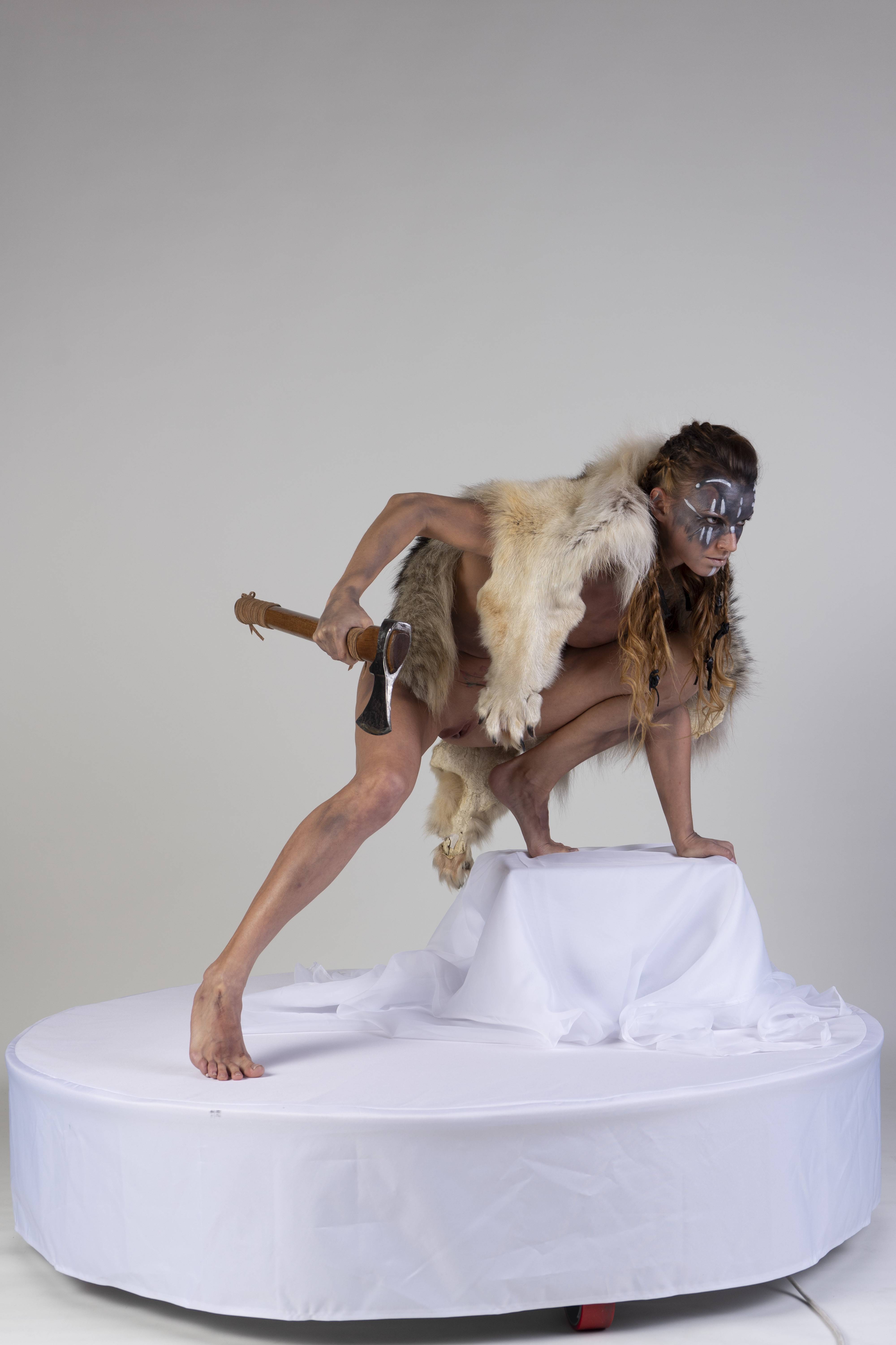 a woman dressed in a warrior costume is holding an ax while standing on a white platform. she has intricate makeup on her face and wears a fur vest. the woman is crouching with one leg extended forward and the other bent, her hands resting on a smaller white platform. the background is a plain studio backdrop.