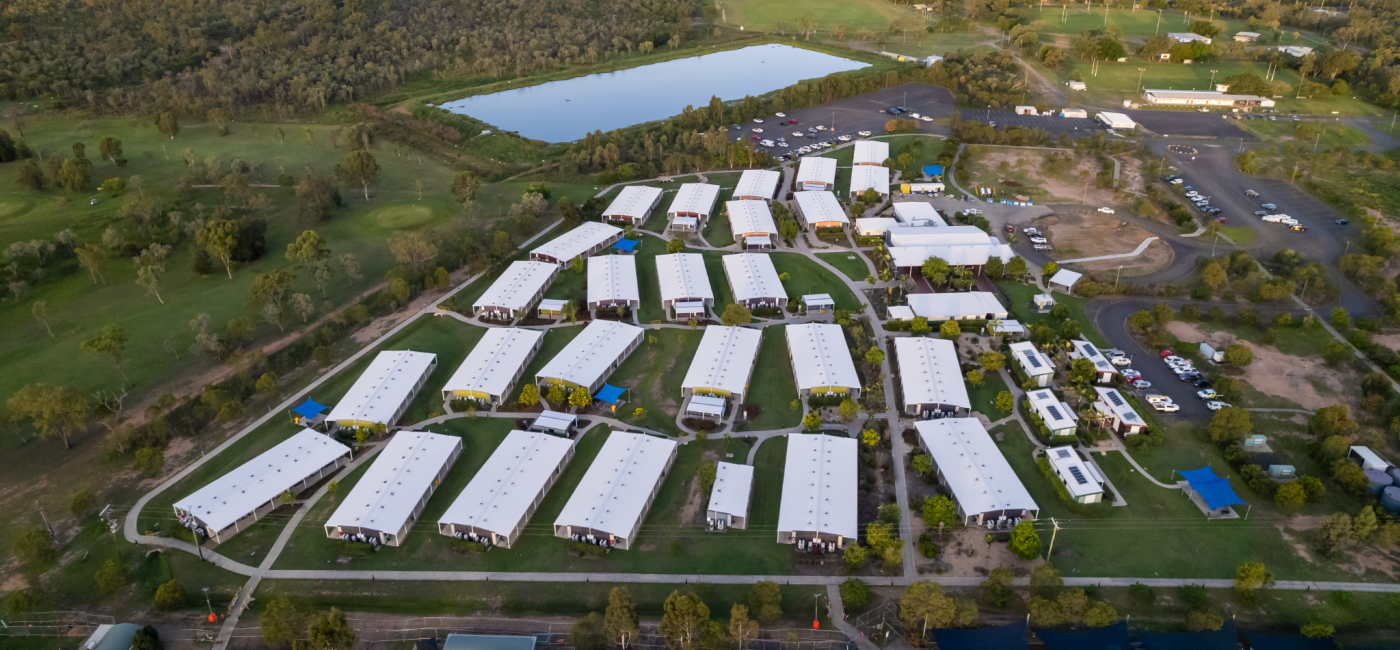 a wide aerial photograph of a suburban area, with many rows of identical, whiteroofed buildings set on a large plot of green grass and parkland. a pond or lake is in the top left of the image. a small parking lot with a few cars is in the top right of the image. there are trees scattered throughout the property. the overall look is a very clean and organized suburb.