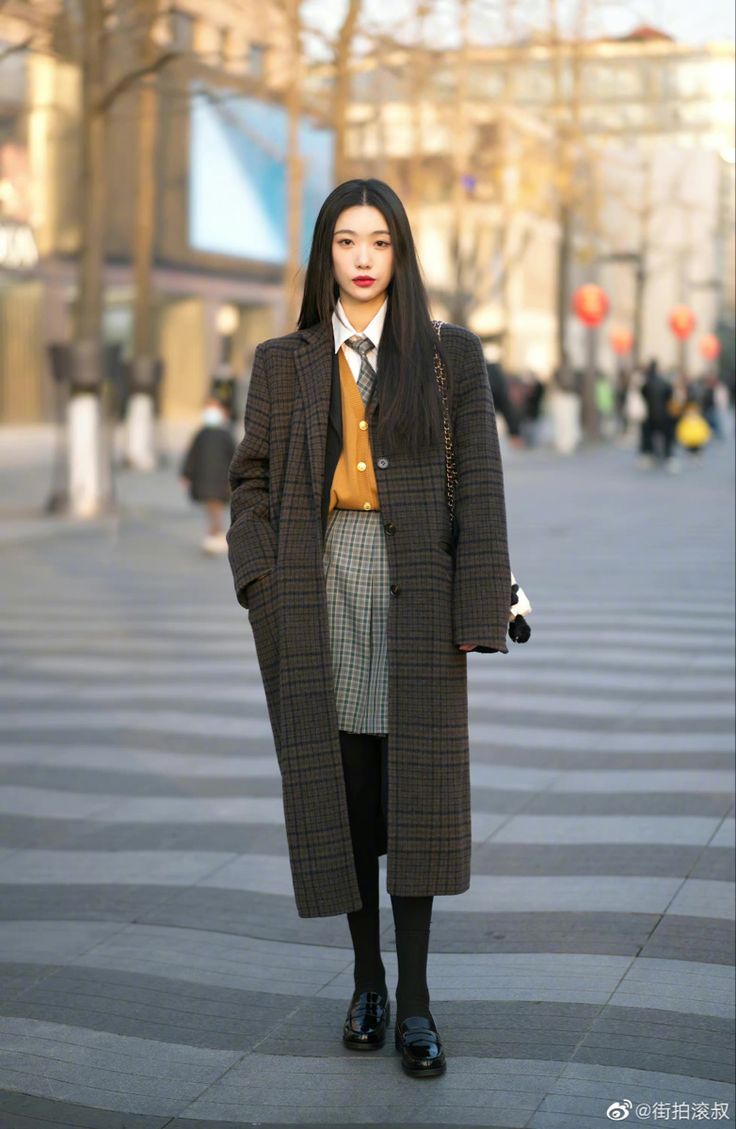 a stylish young woman strolls down a city street. she's dressed in a long brown plaid overcoat, a mustard yellow sweater vest, a pleated gray skirt, black tights and black loafers.  she carries a black handbag over her shoulder. the setting appears to be autumn or winter, judging by the woman's attire and the bare trees in the background. the overall style is elegant and chic, reflecting current street style trends.