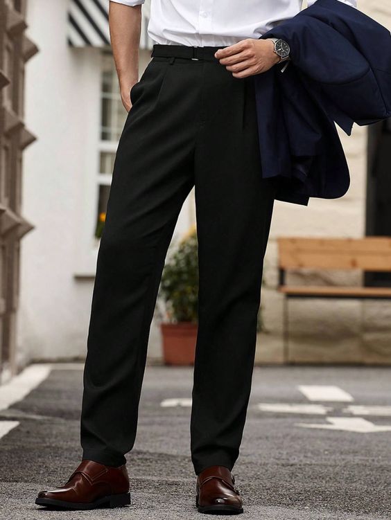 a man is wearing a black suit with a white shirt and brown shoes. he is standing on a sidewalk in an urban setting. the man is looking down at the ground and has a serious expression on his face. he is dressed in a stylish and professional manner.
