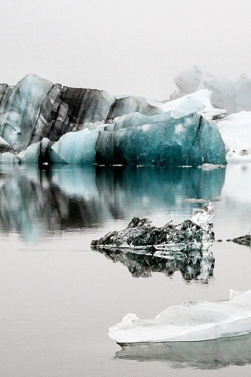 the image depicts a glacier lagoon in iceland, featuring large icebergs floating in calm water. the icebergs are various shades of blue and white, and their reflections are visible in the water. the landscape is cold and serene, with a wintery feel.