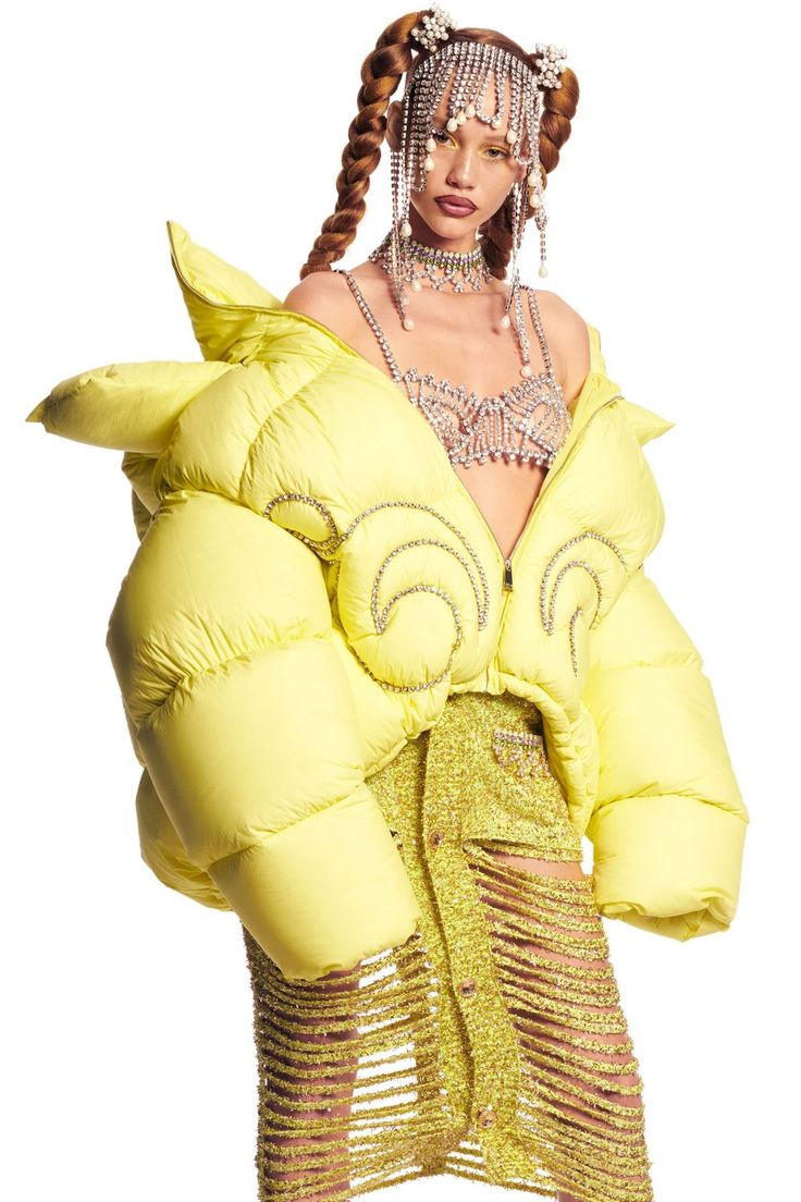 a young woman with long, blonde hair and a jeweled hairpiece stands in a studio with a white background. she is wearing a yellow, puffy jacket with embellished detailing on the front. she is wearing a matching yellow,  long, seethrough skirt  and a rhinestone bra top, with a rhinestone choker. she is posing with her hands on her hips.