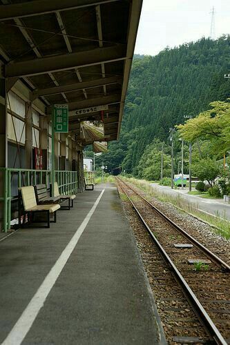 this is an image of a rural japanese train station. the station is small and simple, with a few benches and a green roof. there are train tracks in the foreground and mountains in the background. the sky is overcast and the overall atmosphere is serene and peaceful.