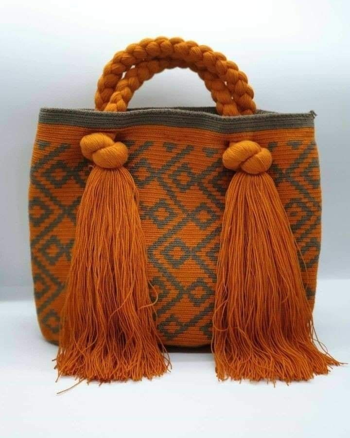 a knitted orange and gray tote bag with tassels. the bag has a woven pattern with a geometric design. the tassels are made of the same material as the bag and are attached to the top handles. the bag is a perfect accessory for any casual occasion.