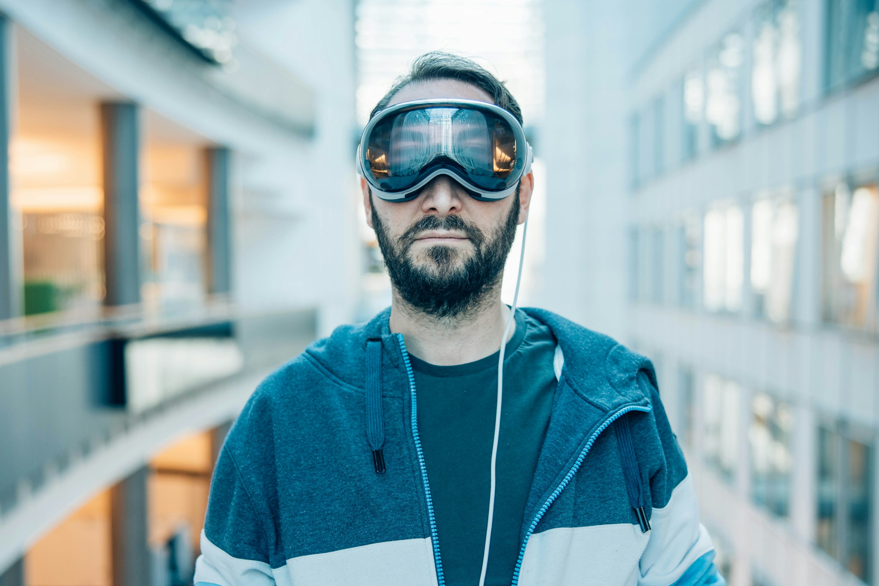 a man with a beard stands indoors wearing virtual reality goggles. he is wearing a twotone hoodie and a tshirt. a white cable is connected to the goggles. the background shows a modern building interior with windows and structures.
