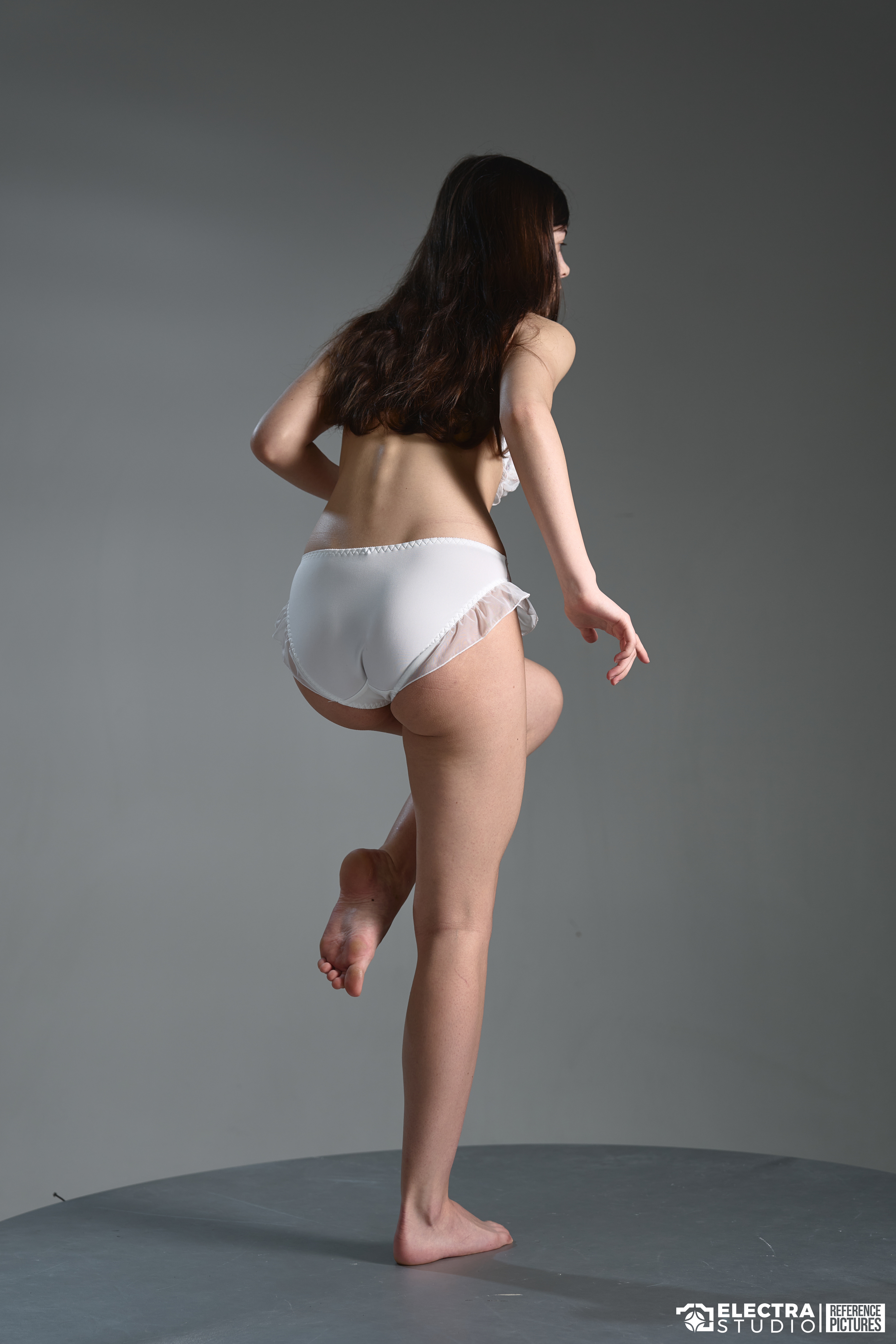 a studio shot of a woman in white lingerie, seen from the back, standing on one leg with the other leg raised. she has brown hair and is barefoot.