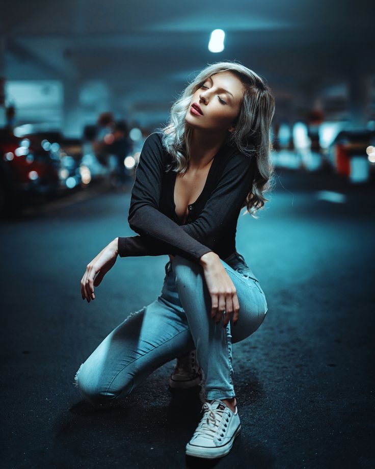 a woman is sitting with her legs crossed in an urban setting at night. she is wearing blue jeans, black shirt, and white sneakers. she has blonde hair and is looking up with her eyes closed. the image is lit with warm and cool lighting, creating a moody and stylish atmosphere.