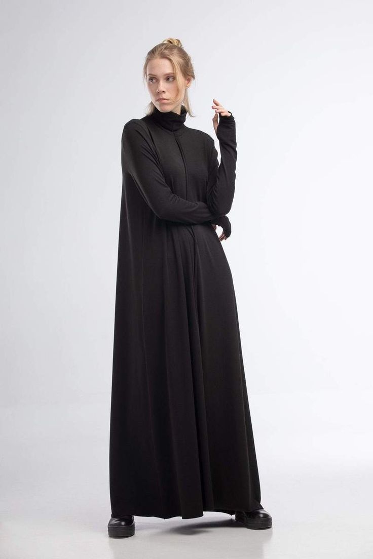 a fulllength studio shot of a blonde woman wearing a black maxi dress. the dress is a simple, aline style with a high turtleneck and long sleeves. she is posing with one hand near her face. the background is plain white.  she is wearing black shoes.