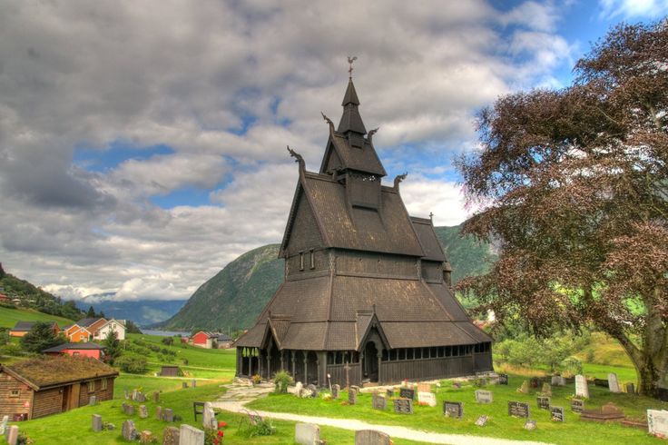 a photograph shows the urnes stave church in norway, sitting atop a grassy hill surrounded by a graveyard, a village, and mountains, set against a cloudy sky. the church is dark brown, constructed of wood, with a tall, layered roof structure culminating in a spire. the graveyard surrounds the church with many headstones scattered around. a small village is in the background, consisting of small houses on the slopes of mountains in the background. a large tree sits to the right, partly obscuring the view. the sky is filled with clouds with patches of blue sky visible.