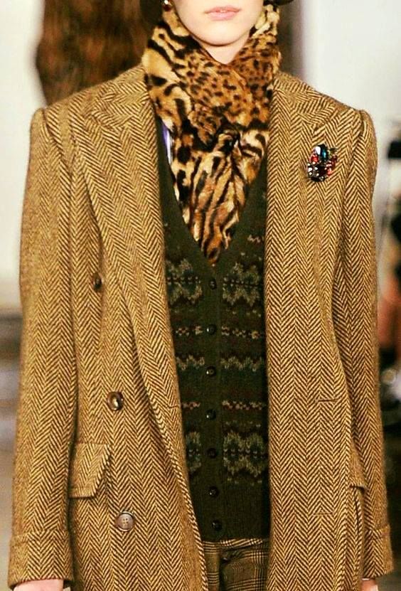 loseup shot of a woman modeling a stylish outfit. the focal point is a gold herringbone tweed doublebreasted jacket.  she's also wearing a leopard print scarf, a dark green fair isle cardigan, and plaid pants. the jacket has a brooch pinned to it. the overall style is reminiscent of vintage or 90s fashion, with an autumnal or winter vibe. the image is welllit and shows good detail of the textures and patterns of the clothing.