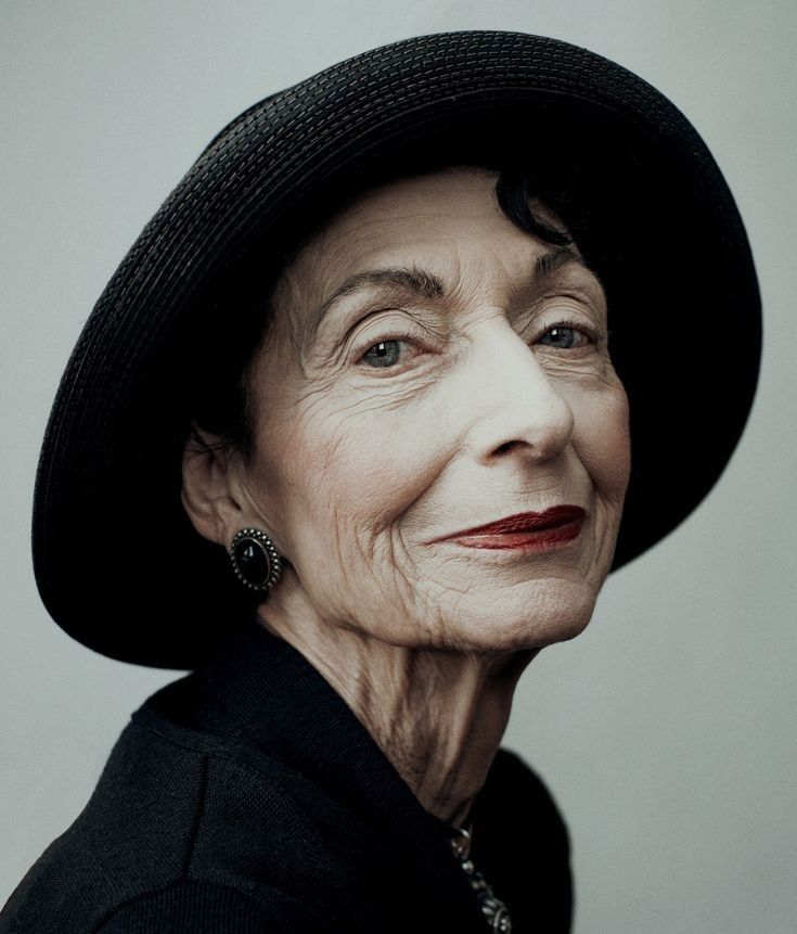 a portrait of an elegant elderly woman wearing a black hat and dark lipstick. she is dressed in black, adorned with jewelry, and displays wrinkles. her expression is serious and confident.
