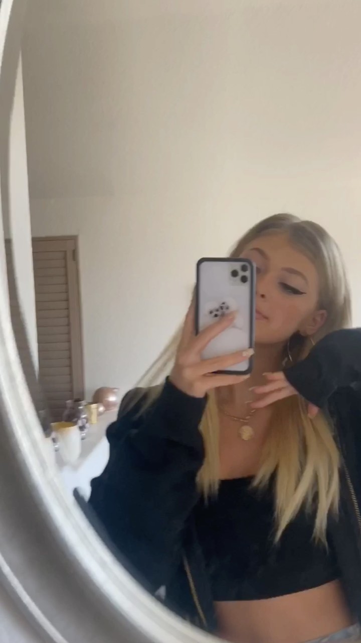 a young woman with blonde hair is taking a mirror selfie with her iphone. she is wearing eyeliner, a necklace, a black top, and a jacket. she is standing indoors in front of a mirror. her facial features are welldefined and her makeup is visible. the background is blurred. she has cosmetic products in the background of the photo.