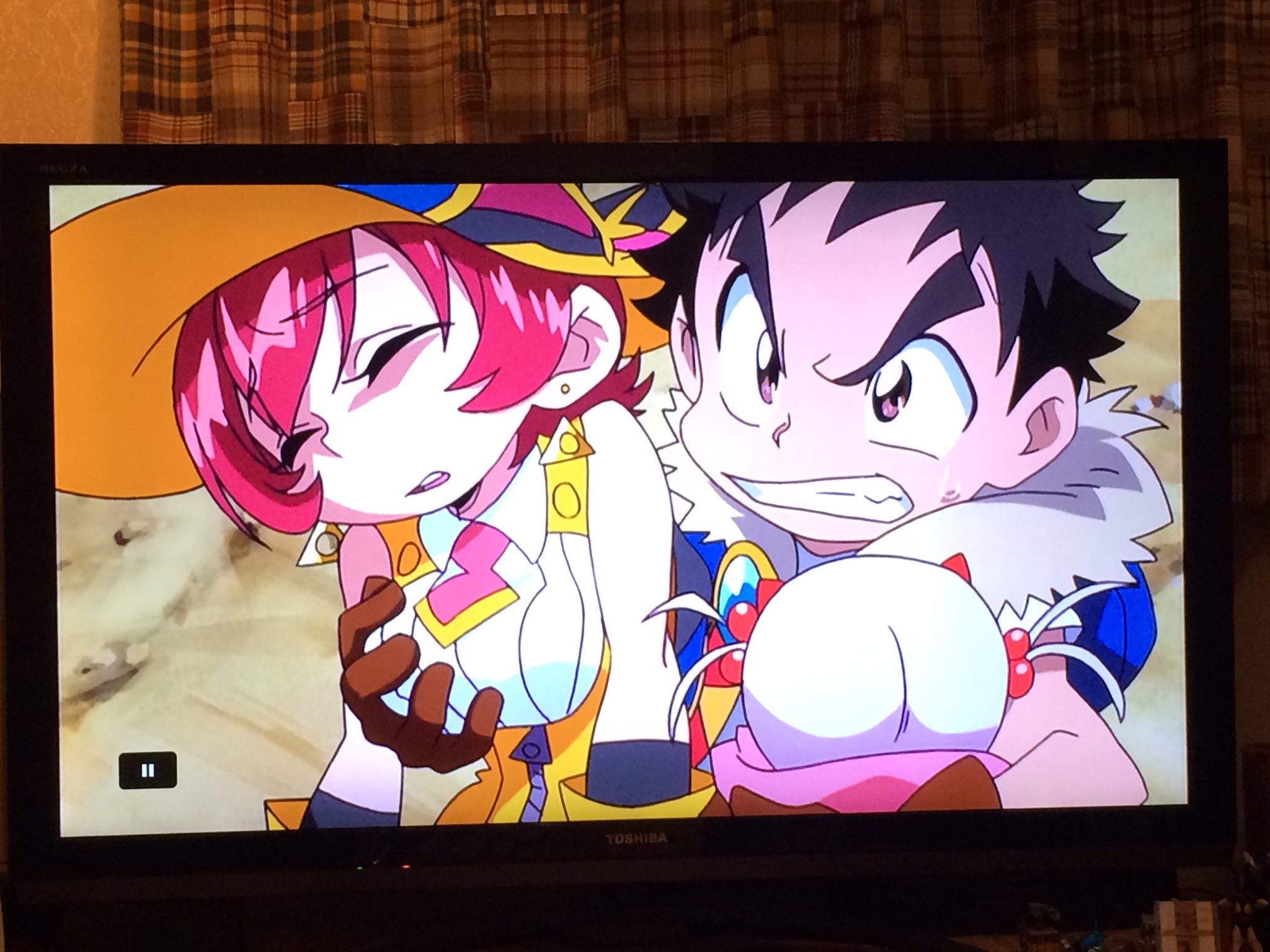a still image from an anime television show displayed on a flatscreen tv. the scene depicts a male and female character in what appears to be a moment of tension or action. the male character, with brown hair and blue eyes, is grasping the female character, who has red hair and is wearing a yellow and white outfit. she appears to be in distress. the background suggests a desert or canyon setting.