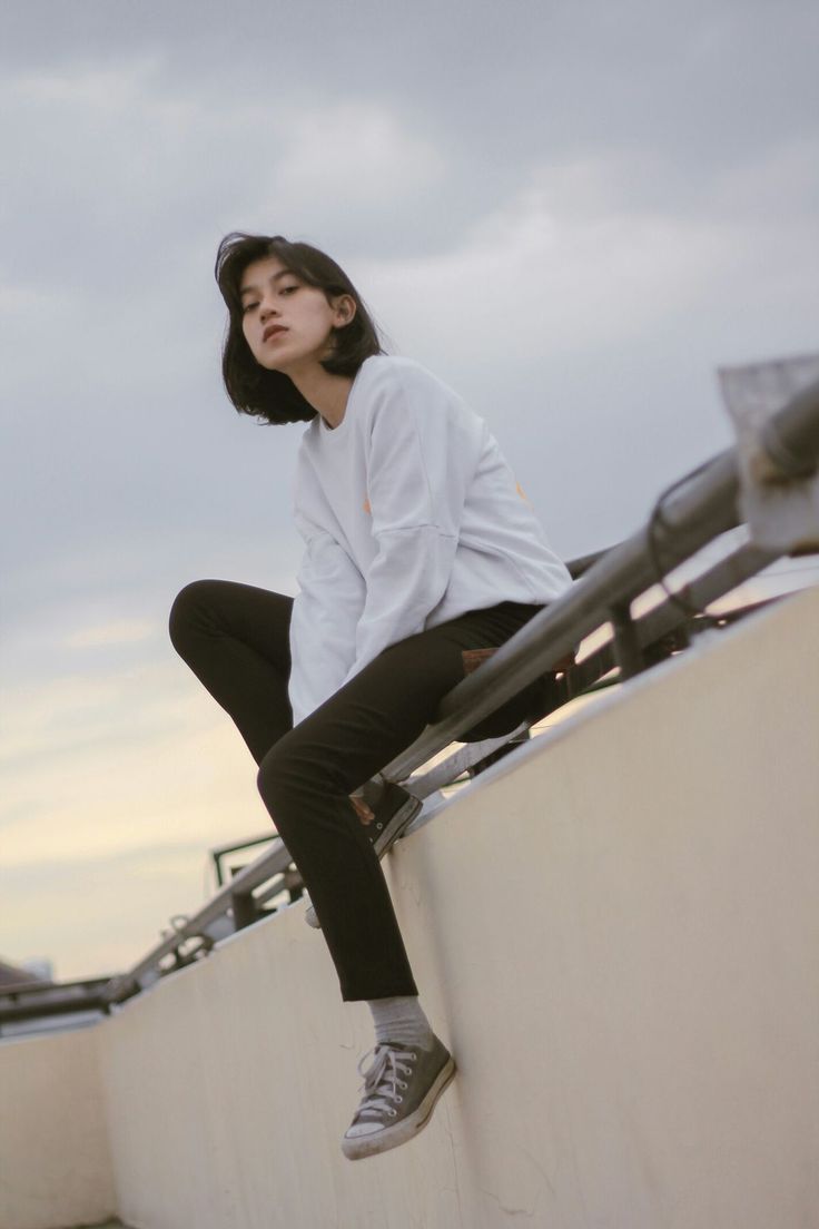 a young woman in a white sweater and black jeans sits on a concrete wall. she is looking at the camera. she has her feet up on the wall and is wearing sneakers. the background is a cloudy sky.