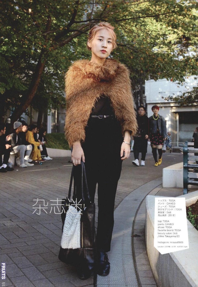 a young woman is posing for a street style photo in tokyo. she's wearing a brown furry shawl, a black top, black pants, and black loafers. she's also carrying a large black and white tote bag. the photo has a caption in japanese that says "fruits 55" and some text that lists the brands of her clothing. the background of the photo shows a street scene with other people and trees.