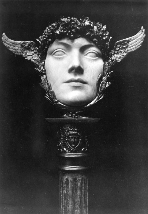 a black and white photograph shows a sculpture of a woman's head on a pedestal. the head is smooth and featureless, adorned with a crown of flowers and winged ornaments on either side of the head. the sculpture is placed on a square platform, which sits atop a more ornate section featuring another face sculpture. the entire structure rests on a fluted column. the background is a dark, solid color, which gives the artwork a dramatic and timeless quality.