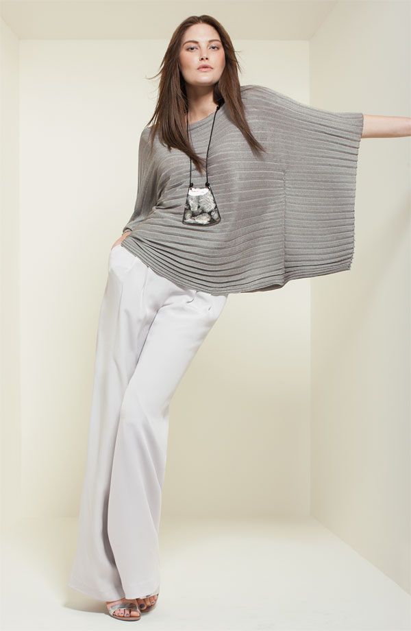 a woman is posing in a studio wearing a gray top, white pants, and sandals. she is wearing a necklace and leaning against a wall.