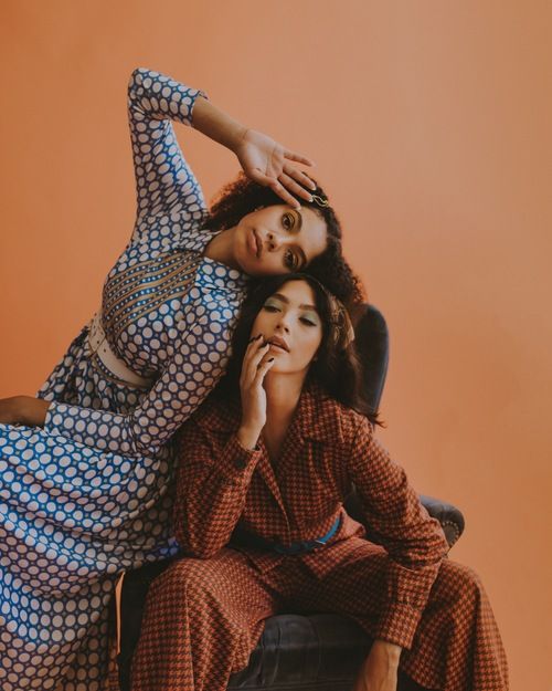 two women are posing in a studio. the woman on the left is wearing a vintage patterned dress and is leaning towards the right. she has her arm raised over the head of the woman sitting to her right. the woman on the right is wearing an orange patterned jumpsuit and is looking directly at the camera. both women are wearing light makeup. they both appear to be friends enjoying themselves.