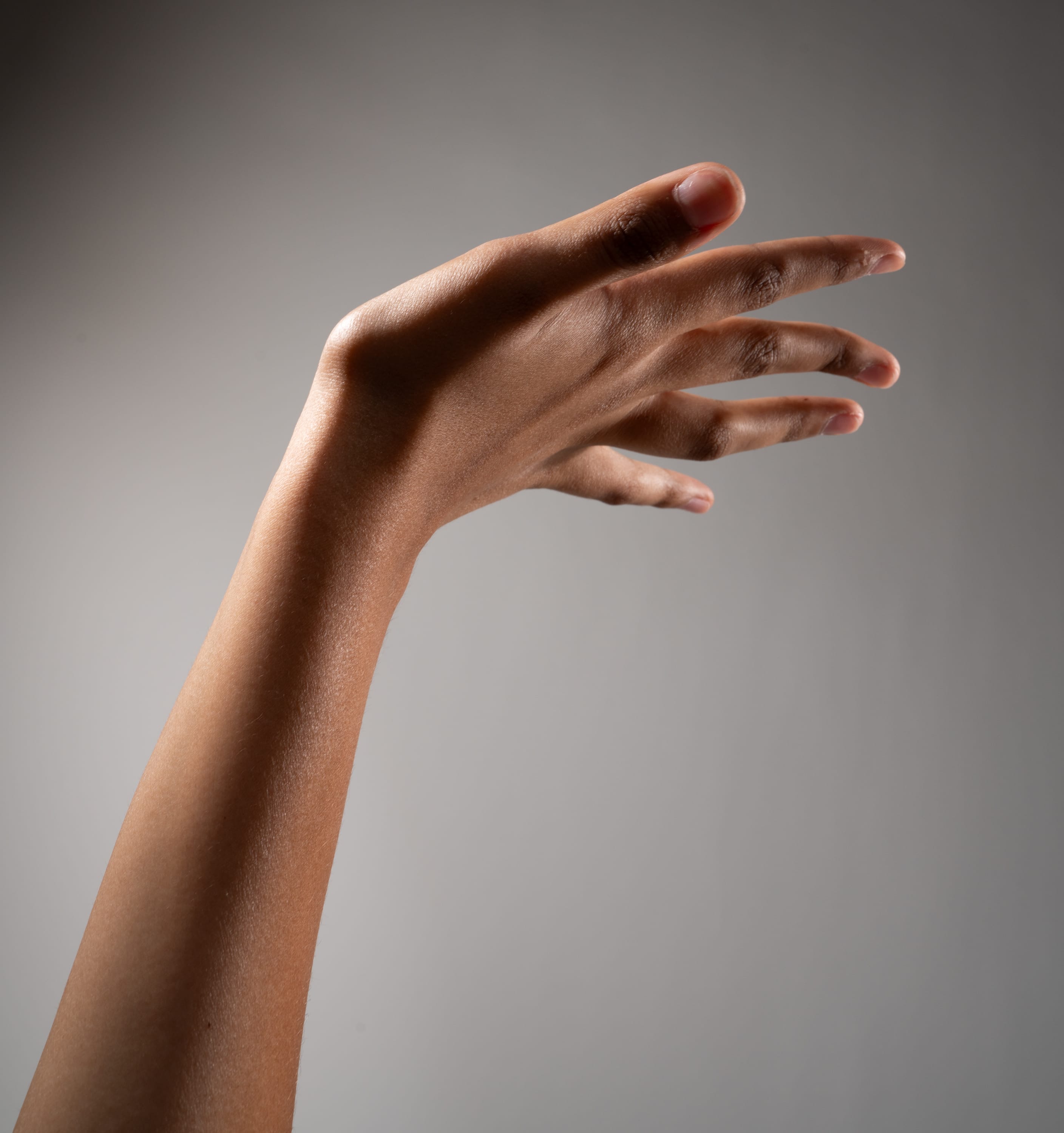 the image shows a hand and arm of a person with a dark skin tone extended against a neutral gray background. the hand is held up, fingers outstretched, and is illuminated with soft lighting, creating a natural and subtle shadow on the arm. the image focuses solely on the hand and arm, highlighting the details of its form and skin tone.