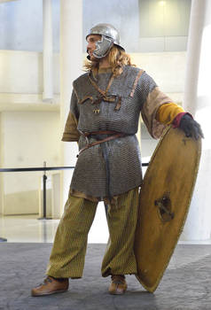 a man dressed as a roman soldier stands in a museum. he is wearing a chainmail shirt, a helmet, and trousers. he also carries a wooden shield. the setting appears to be indoors, with a modern architectural backdrop. the overall impression is that of a historical reenactment or museum display.
