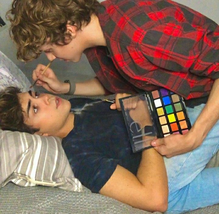 a young man is applying eyeshadow to another young man's eye with a brush. they are laying on a bed and are smiling and looking at each other. they are both wearing casual clothing. the room is decorated with a colorful eyeshadow palette on the bed. it appears they are in a playful and affectionate mood.