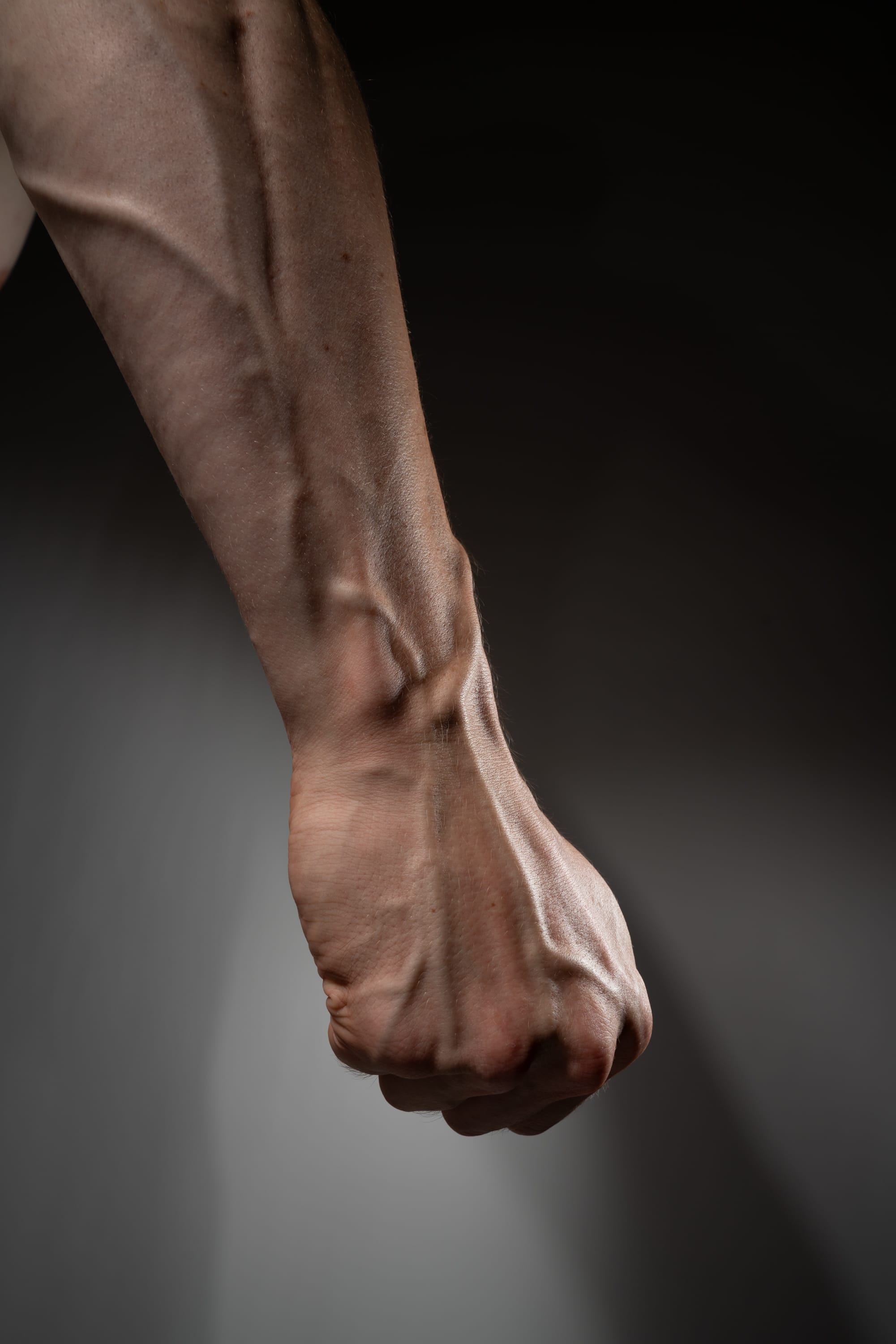 a clenched fist of a man's arm is shown, highlighting the prominent veins and muscular structure against a dark background. the image evokes themes of strength, power, and tension. the lighting emphasizes the contours of the arm, creating a dramatic effect.