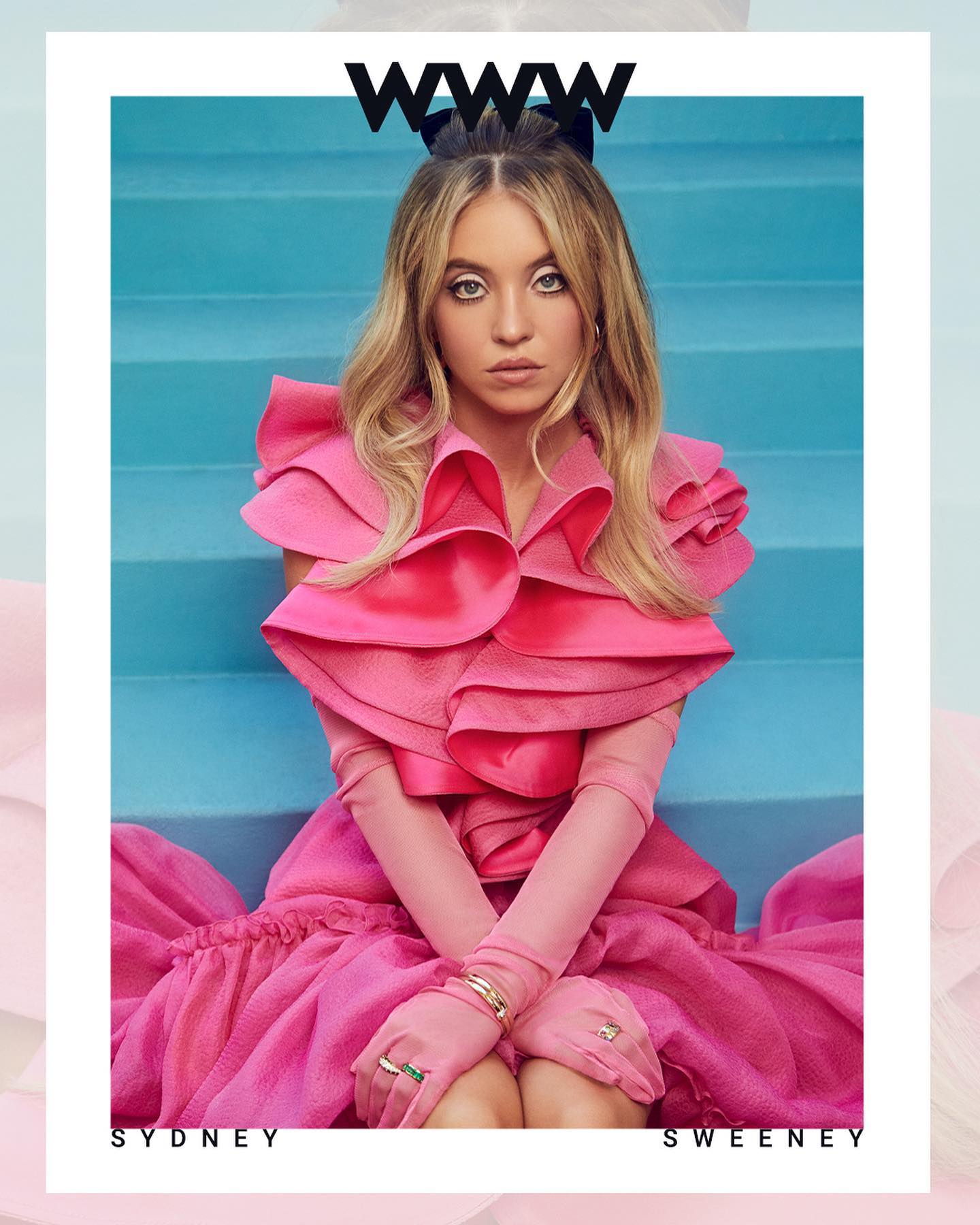 the image is a magazine cover featuring actress sydney sweeney. she is centrally positioned and seated on what appears to be aquacolored stairs. sweeney wears a vibrant, hot pink, ruffled dress with long pink gloves.  she's accessorized with several rings. her blonde hair is styled neatly. the background is a simple, yet visually appealing backdrop.  the "www" magazine logo is prominent at the top. the text "sydney sweeney" is subtly placed at the bottom.  the overall aesthetic is stylish and glamorous.