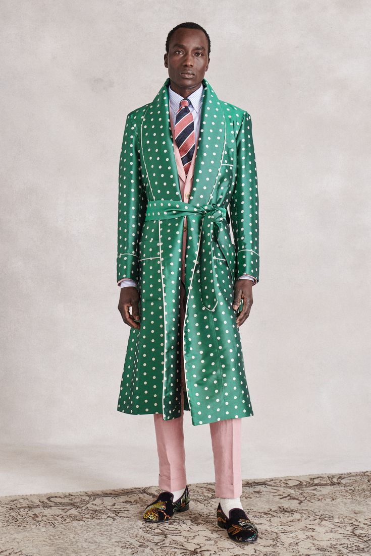 a fulllength studio shot of a male fashion model showcasing a green silk polka dot robe. the robe has a shawl collar, a tie waist, and is detailed with white piping. the model is wearing a pink shirt and trousers underneath the robe and is wearing embroidered loafers. the background is a neutral, light gray. the overall style is formal and elegant.