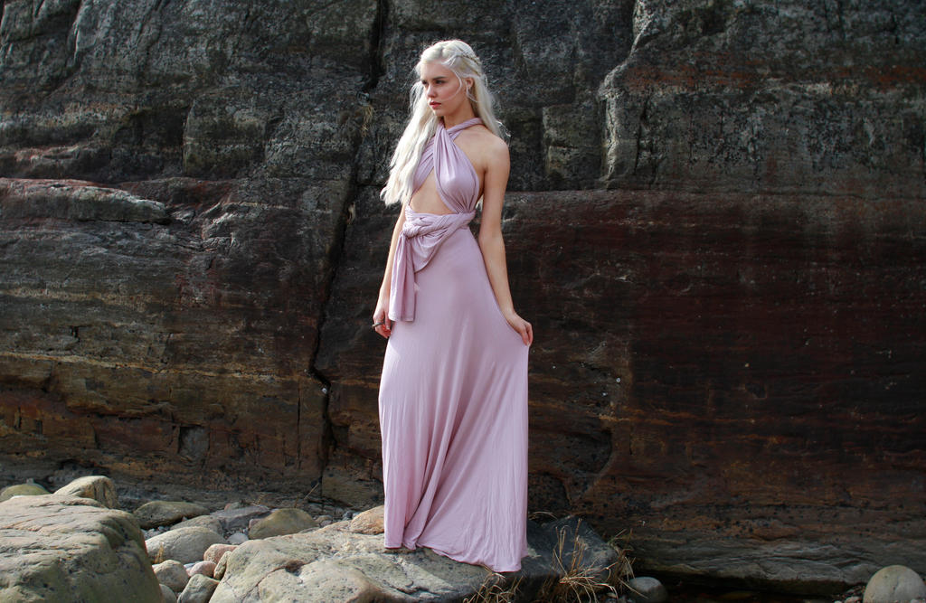 a fulllength shot of a blonde woman standing on a rocky beach, wearing a long, flowing pink infinity dress. the dress is styled in a halter neck and wrap design, revealing a subtle midriff.  her hair is styled in a loose braid. the background is a large, darkcolored rock formation. the overall mood is serene and somewhat mystical.