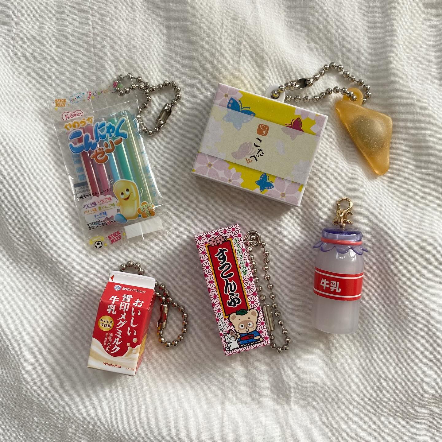 the image shows a flat lay of several keychains featuring japanese snacks and products. there are keychains of konjac jelly sticks, a milk carton, a small box with japanese designs, kombu seaweed, and a milk bottle. they are all arranged on a white wrinkled cloth.