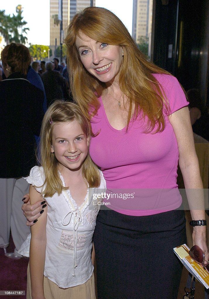 a woman with long red hair and a pink shirt, is smiling next to a young girl. the girl is wearing a white shirt and has long blonde hair. both are smiling happily as they pose for a photo together. the woman has her arm around the girl. they are outside on a street.