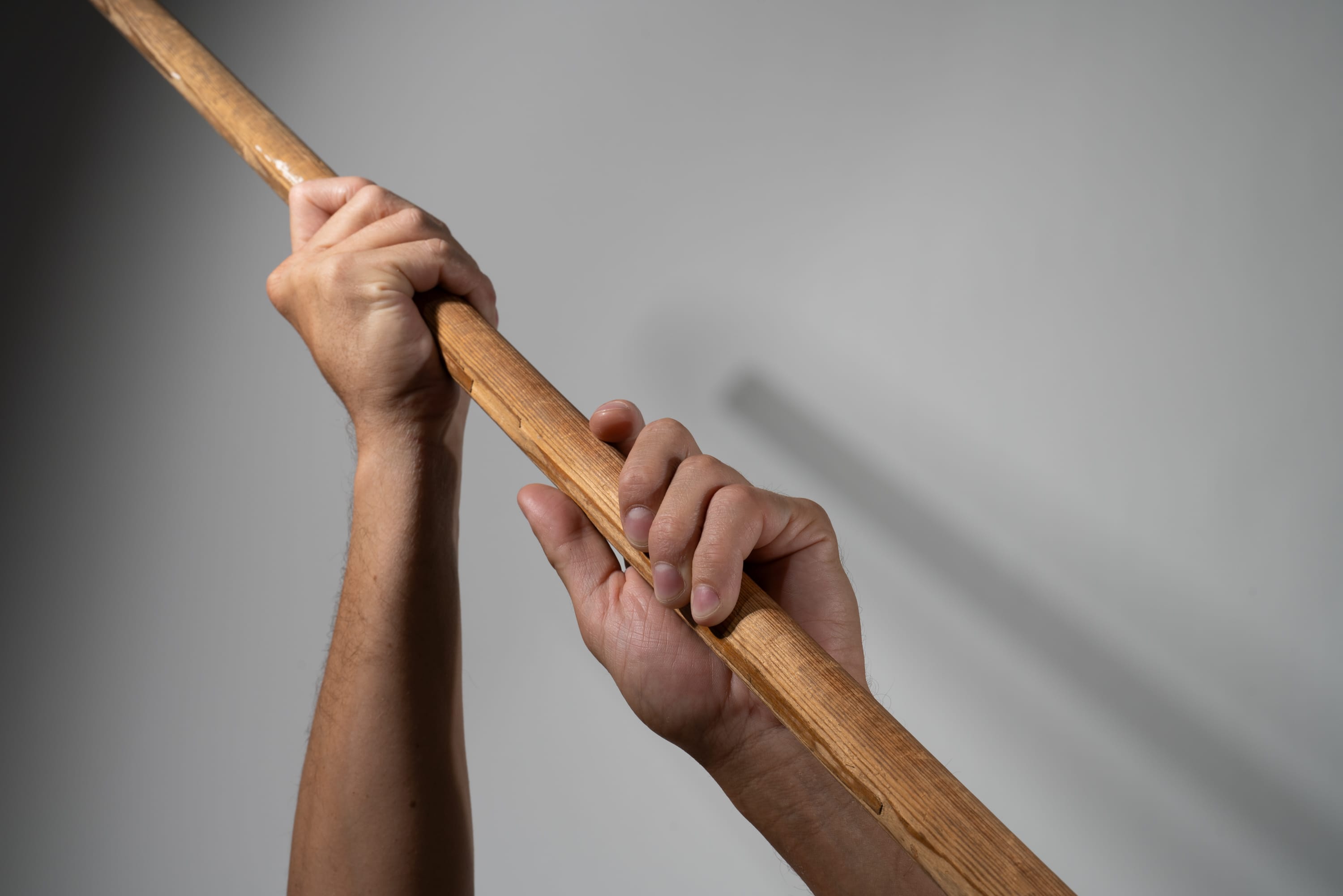 two hands grip a wooden pole against a plain, light gray background. the perspective is from below, looking up at the hands and the pole, which extends out of frame. the image is brightly lit, creating soft shadows on the wall behind.