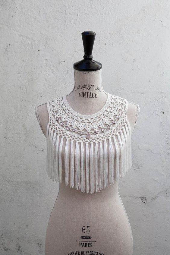 a white crochet bib necklace with fringe detailing is displayed on a vintage mannequin. the necklace features intricate crochet work with small beads and long tassels. the mannequin is creamcolored and stands against a white textured background. the image is lit with soft, natural light.